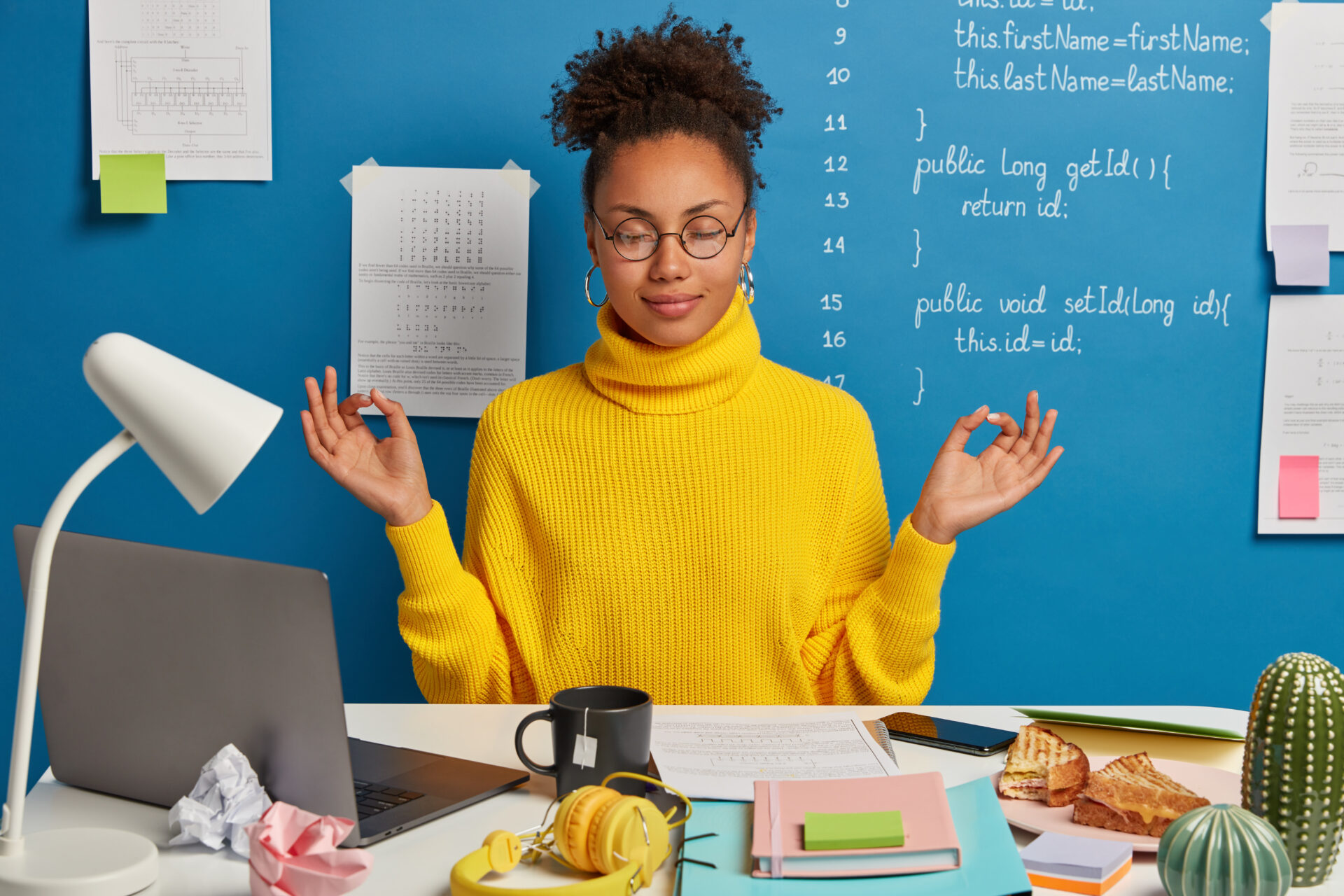 Woman freelance worker does yoga exersice at workplace, enjoys calm tranquil atmosphere, wears round glasses and jumper, gathers with thoughts, takes break after working on laptop and with papers
